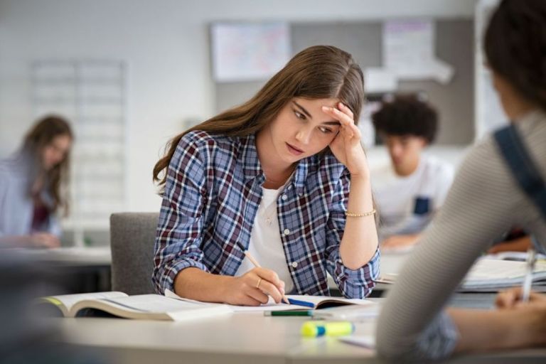 Anxious young woman with hand on head feeling tired while studying at school in classroom. College student suffering from headache in library preparing for test. Portrait of troubled and stressed girl doing exam that doesn